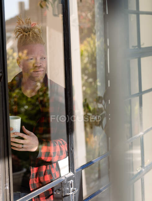 Thoughtful albino african american man with dreadlocks drinking coffee. remote working using technology at home. — Stock Photo