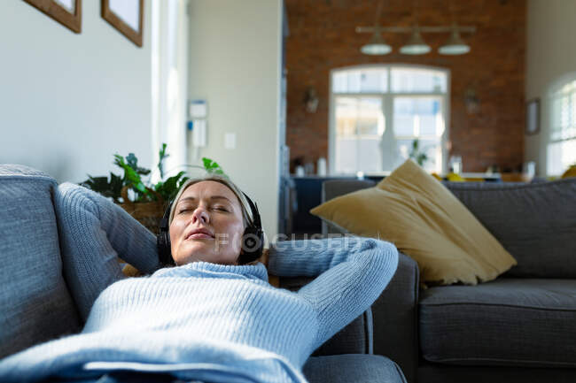 Розслаблена старша біла жінка у вітальні лежить на дивані, одягнена в навушники. пенсійний спосіб життя, вдома з технологіями . — стокове фото