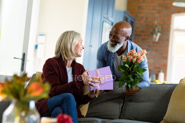 Щаслива старша різноманітна пара у вітальні сидить на дивані, даруючи квіти та подарунки. пенсійний спосіб життя, проводити час вдома . — стокове фото