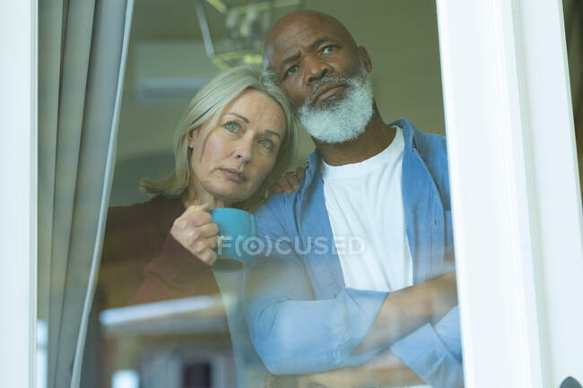 Worried senior diverse couple looking through window and embracing. retirement lifestyle, spending time at home. — Stock Photo