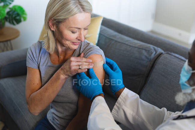 Fröhliche Seniorin und Ärztin mit Mundschutz sitzen im Wohnzimmer auf dem Sofa und impfen. Gesundheit und Lebensstil von Senioren während der covid 19 Pandemie. — Stockfoto