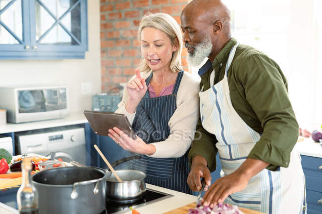 Happy senior diverse couple in kitchen wearing aprons, cooking together, using tablet. healthy, active retirement lifestyle at home. — Stock Photo
