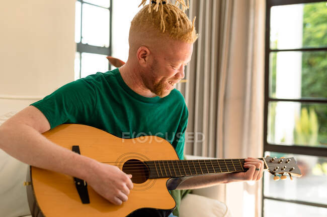 Улыбающийся американский альбинос с дредами в гостиной, играющий на гитаре. свободное время, отдых дома. — стоковое фото