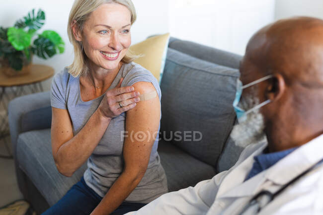 Счастливая старшая разнообразная женщина и врач в масках для лица в гостиной сидит на диване, вакцинируя. здоровье и образ жизни пожилых людей во время пандемии ковида 19. — стоковое фото