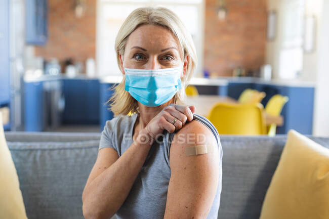 Glückliche ältere kaukasische Frau mit Gesichtsmaske im Wohnzimmer mit Verband am Arm. Gesundheit und Lebensstil von Senioren während der covid 19 Pandemie. — Stockfoto