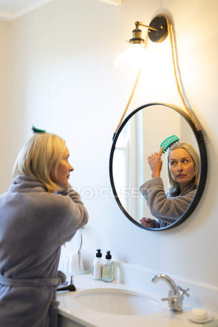 Mulher caucasiana sênior feliz no banheiro, olhando para o espelho, escovando o cabelo. estilo de vida da aposentadoria, passar tempo em casa. — Fotografia de Stock