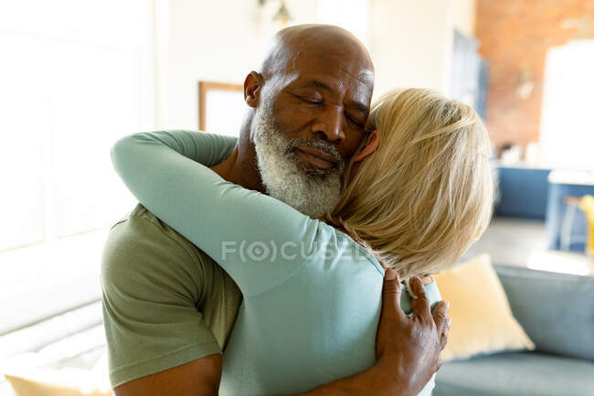 Feliz pareja de ancianos diversos en la sala de estar abrazando con los ojos cerrados. estilo de vida de jubilación, pasar tiempo en casa. - foto de stock
