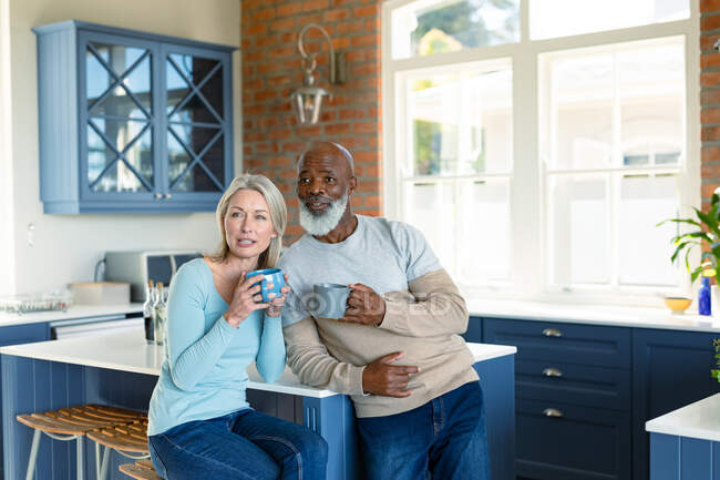 Happy senior diverse couple in kitchen sitting at countertop, drinking coffee. retirement lifestyle, spending time at home. — Stock Photo
