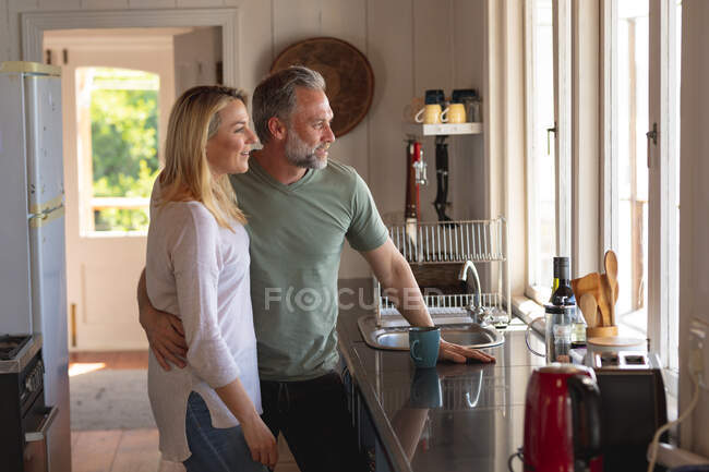 Happy caucasian mature couple drinking coffee in the kitchen and looking through the window. enjoying leisure time at home. — Stock Photo
