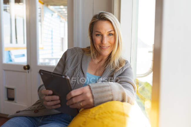 Glückliche kaukasische reife Frau mit Tablette im sonnigen Wohnzimmer. Freizeit zu Hause genießen. — Stockfoto