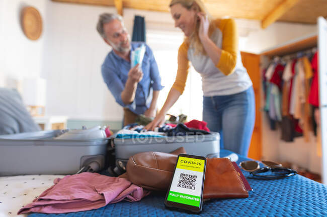 Smartphone mit covid 19 Impfpass, im Hintergrund ein kaukasisches Paar beim Kofferpacken. Reise- und Urlaubsvorbereitungen während der covid 19 Pandemie. — Stockfoto