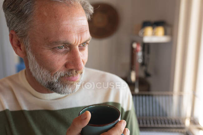 Rilassante uomo maturo caucasico che beve caffè in cucina e guarda attraverso la finestra. godendo del tempo libero a casa. — Foto stock