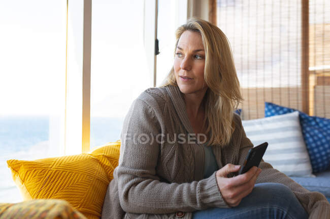 Kaukasische reife Frau mit Smartphone und Blick durchs Wohnzimmerfenster. Freizeit zu Hause genießen. — Stockfoto