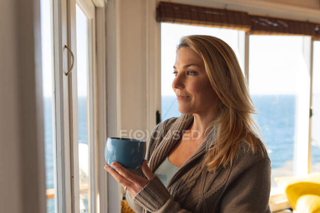 Rilassante donna matura caucasica bere caffè in cucina e guardare attraverso la finestra. godendo del tempo libero a casa. — Foto stock