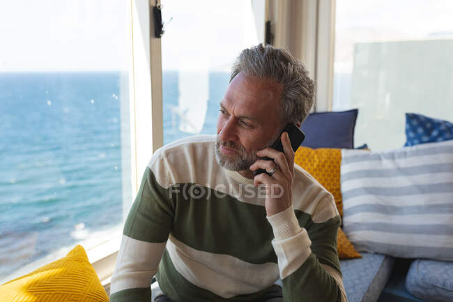 Kaukasischer reifer Mann mit Smartphone und Blick durchs Wohnzimmerfenster. Freizeit zu Hause genießen. — Stockfoto