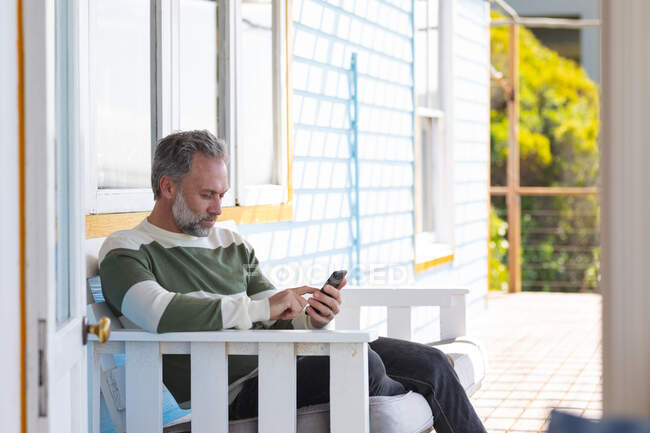Caucásico hombre maduro utilizando el teléfono inteligente en una terraza junto al mar. disfrutar del tiempo libre en la casa frente a la playa. - foto de stock