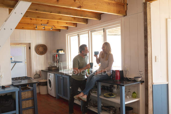 Счастливая кавказская взрослая пара пьет кофе на солнечной кухне. наслаждаясь отдыхом дома. — стоковое фото