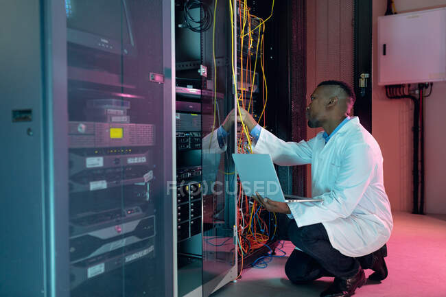 Técnico de computación masculino afroamericano que usa computadora portátil que trabaja en la sala de servidores de negocios. tecnología de redes digitales de almacenamiento y comunicación de información. - foto de stock