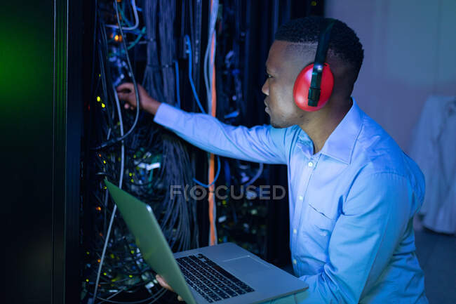 Técnico de computadoras afroamericano usando auriculares usando computadora portátil trabajando en la sala de servidores. tecnología de redes digitales de almacenamiento y comunicación de información. - foto de stock