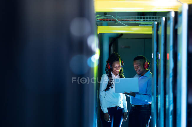 Techniciens informatiques afro-américains portant des écouteurs utilisant un ordinateur portable travaillant dans la salle des serveurs. stockage de l'information numérique et technologie des réseaux de communication. — Photo de stock