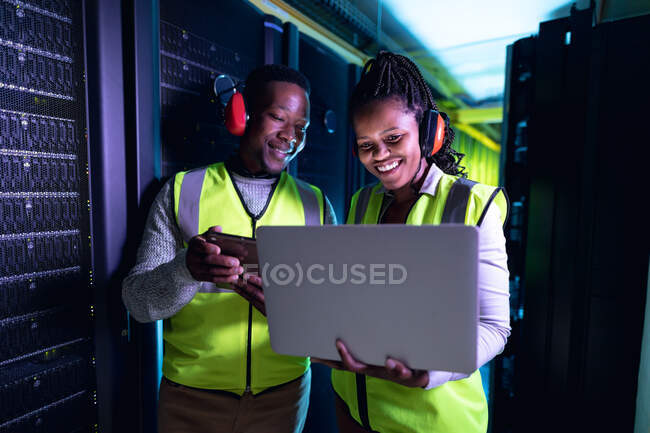 Técnicos africanos americanos felices usando auriculares usando el ordenador portátil que trabaja en la sala de servidores. tecnología de redes digitales de almacenamiento y comunicación de información. - foto de stock