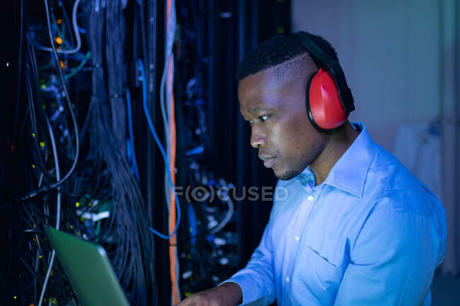 Африканский американец, компьютерный техник, носит наушники, используя ноутбук, работающий в серверной. цифровые технологии хранения и передачи информации. — стоковое фото