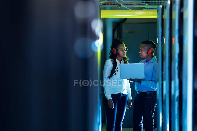 Técnicos de informática afro-americanos usando fones de ouvido usando laptop trabalhando na sala do servidor. armazenamento digital de informações e tecnologia de rede de comunicação. — Fotografia de Stock