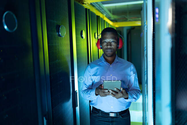 Africano americano técnico de computador do sexo masculino usando fones de ouvido usando tablet trabalhando na sala do servidor. armazenamento digital de informações e tecnologia de rede de comunicação. — Fotografia de Stock