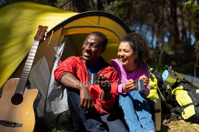 Happy diverse couple sitting in the tent and drinking coffee in countryside. healthy, active outdoor lifestyle and leisure time. — Stock Photo