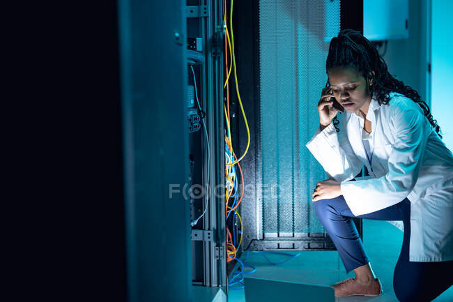 Técnica de informática afro-americana fazendo chamadas e usando laptop trabalhando na sala de servidores. armazenamento digital de informações e tecnologia de rede de comunicação. — Fotografia de Stock