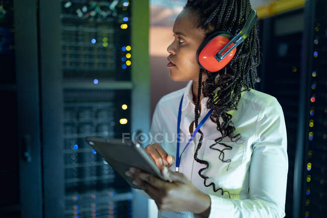 African american female computer technician wearing headphones using tablet working in server room. digital information storage and communication network technology. — Stock Photo