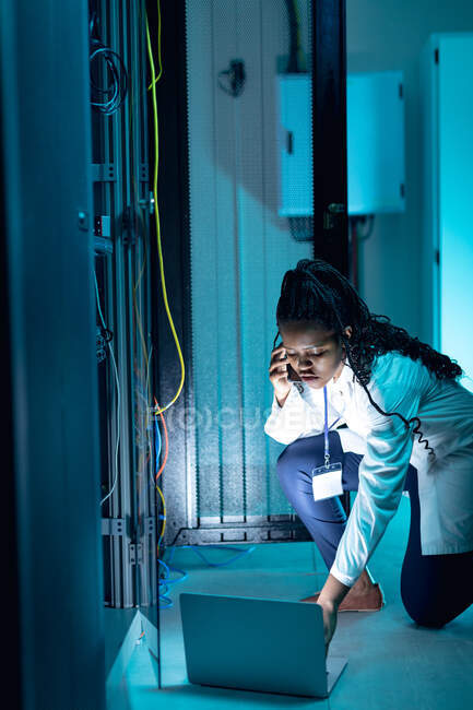 African american female computer technician making call and using laptop working in server room. digital information storage and communication network technology. — Stock Photo