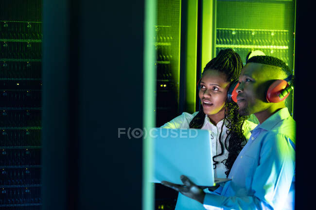 Técnicos informáticos afroamericanos que usan auriculares usando computadoras portátiles que trabajan en la sala de servidores. tecnología de redes digitales de almacenamiento y comunicación de información. - foto de stock