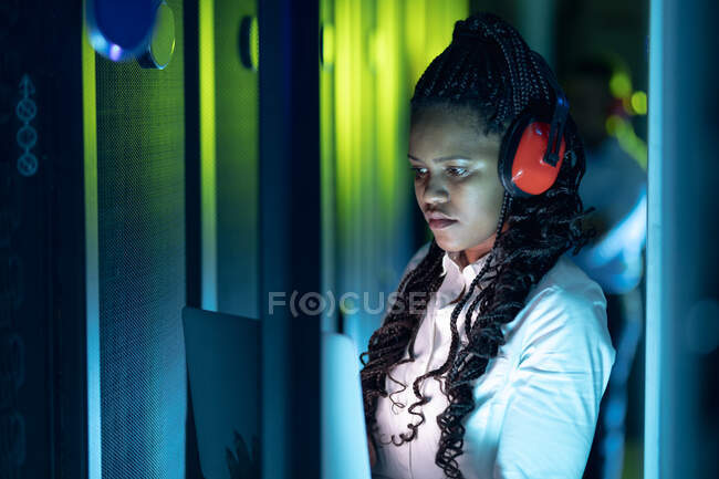 Técnica en computación afroamericana usando auriculares usando tableta trabajando en la sala de servidores. tecnología de redes digitales de almacenamiento y comunicación de información. - foto de stock