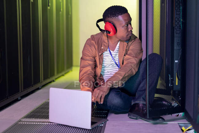 Africano americano técnico de computador masculino usando fones de ouvido usando laptop trabalhando na sala do servidor. armazenamento digital de informações e tecnologia de rede de comunicação. — Fotografia de Stock