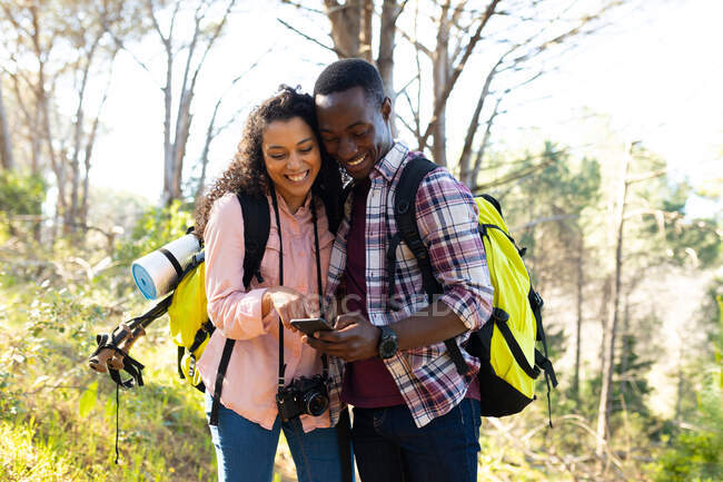 Happy diverse couple with backpacks using smartphone hiking in countryside. healthy, active outdoor lifestyle and leisure time. — Stock Photo