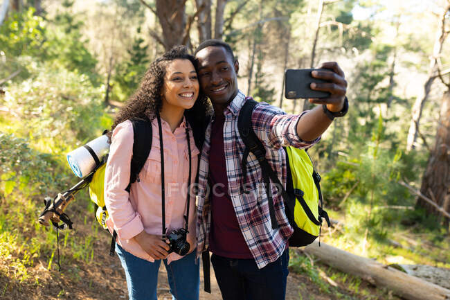 Heureux couple diversifié avec des sacs à dos prenant selfie et randonnée à la campagne. mode de vie sain et actif en plein air et temps libre. — Photo de stock
