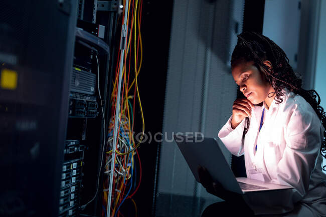 Técnico de computación afroamericana haciendo llamadas y usando laptop trabajando en la sala de servidores. tecnología de redes digitales de almacenamiento y comunicación de información. - foto de stock