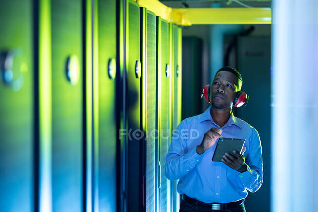 Técnico de computadoras afroamericano usando auriculares usando tableta trabajando en la sala de servidores. tecnología de redes digitales de almacenamiento y comunicación de información. - foto de stock