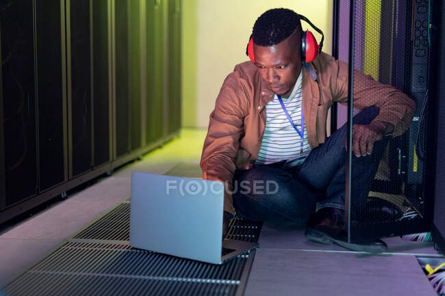 African american male computer technician wearing headphones using laptop working in server room. digital information storage and communication network technology. — Stock Photo
