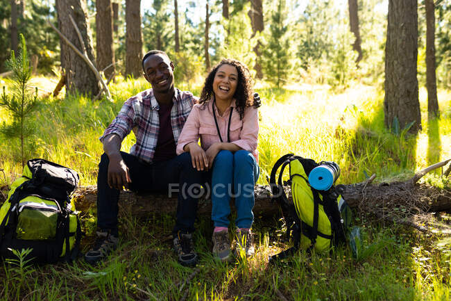 Feliz pareja diversa con mochilas tomando un descanso de senderismo en el campo. estilo de vida al aire libre saludable y activo y tiempo libre. - foto de stock