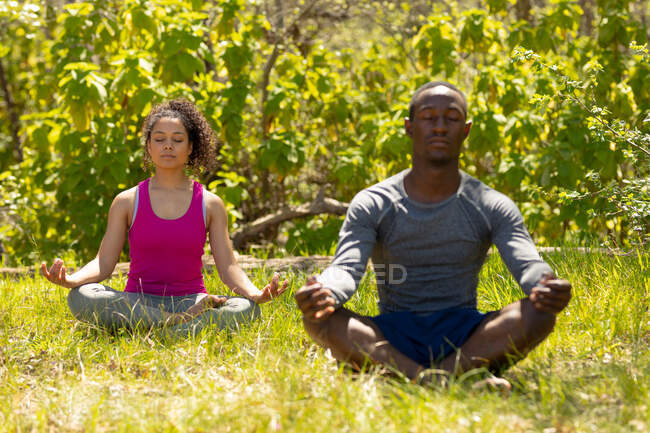 Relaxante casal diversificado sentado com pernas cruzadas e meditando no campo. saudável, estilo de vida ao ar livre ativo e tempo de lazer. — Fotografia de Stock