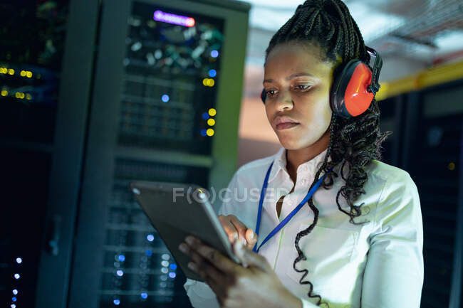 Técnica de informática afro-americana usando fones de ouvido usando tablet trabalhando na sala de servidores. armazenamento digital de informações e tecnologia de rede de comunicação. — Fotografia de Stock