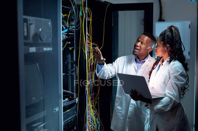 Técnicos informáticos afroamericanos que usan computadoras portátiles que trabajan en la sala de servidores. tecnología de redes digitales de almacenamiento y comunicación de información. - foto de stock