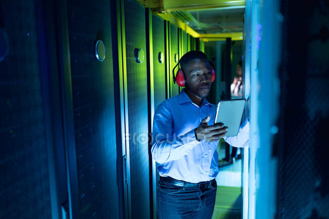 African american male computer technician wearing headphones using tablet working in server room. digital information storage and communication network technology. — Stock Photo