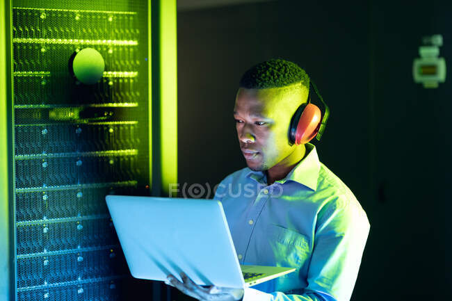 Afrikanisch-amerikanischer Computertechniker mit Kopfhörern und Laptop, der im Serverraum arbeitet. digitale Informationsspeicherung und Kommunikations-Netzwerktechnologie. — Stockfoto