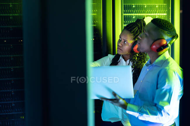 Técnicos informáticos afroamericanos que usan auriculares usando computadoras portátiles que trabajan en la sala de servidores. tecnología de redes digitales de almacenamiento y comunicación de información. - foto de stock