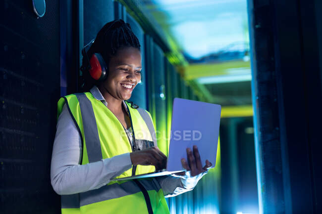 African american female computer technician wearing headphones using laptop working in server room. digital information storage and communication network technology. — Stock Photo