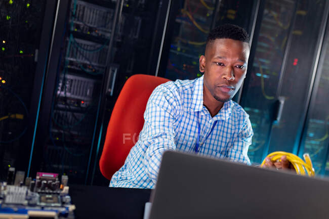 Técnico de computación masculino afroamericano usando portátil que trabaja en la sala de servidores. tecnología de redes digitales de almacenamiento y comunicación de información. - foto de stock