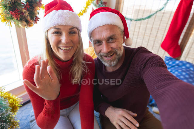 Heureux couple d'âge mûr caucasien portant chapeaux Santa faire un appel vidéo à Noël. Noël, fête et technologie de la communication. — Photo de stock
